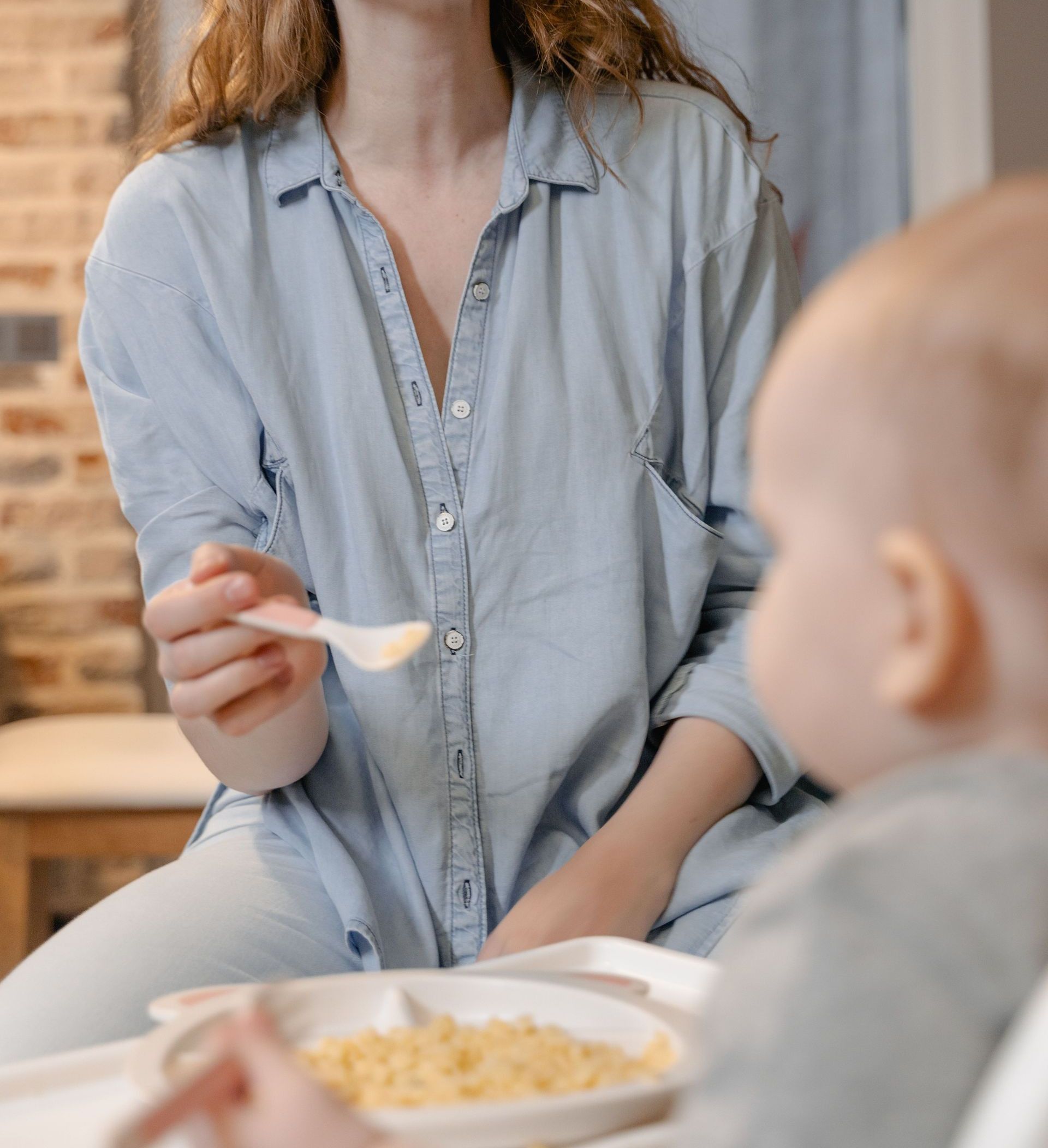A joyful mother feeds her baby a spoonful of pasta indoors, sharing a precious bonding moment.